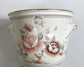 May include: White ceramic planter with a gold rim and handles. The planter is decorated with pink and brown floral designs and blue leaves. The planter has a pedestal base.