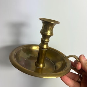 May include: A vintage brass candlestick holder with a round base and a handle. The candlestick has a tall, ornate stem and a flared top to hold a candle. The brass has a warm, golden color.