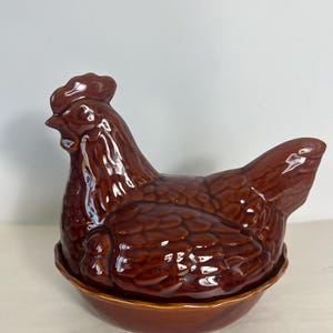Peut inclure: Un plat en céramique marron en forme de poule avec une finition brillante. La poule a une crête et des barbillons détaillés, avec un corps arrondi et une base qui sert de plat. Un article de cuisine décoratif.