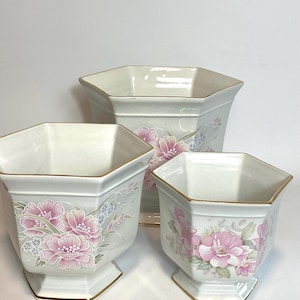 May include: Three white ceramic hexagonal planters with a gold rim and floral designs. The planters are of different sizes and feature pink and blue flowers.