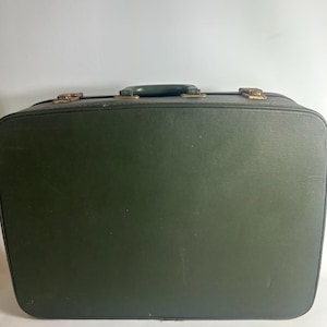 May include: A rectangular dark green suitcase featuring a top handle and two metallic latches. The exterior has a smooth, solid finish with rounded corners and visible wear on the hardware, providing a functional design for travel storage.