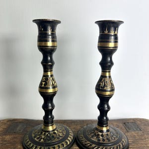 May include: Two antique candle holders in black and gold. The bases feature an etched floral pattern. The bodies are black with gold bands and decorative carvings. The tops are designed to hold candles.