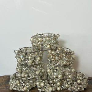 May include: A set of six circular napkin rings constructed from silver-toned wire mesh. Each ring is densely embellished with a variety of clear glass beads and round white faux pearls, creating a textured, multi-dimensional surface appearance.