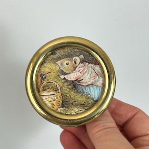 May include: A round, brass-framed miniature painting featuring a mouse in a pink and blue outfit, gathering berries near a woven basket. The artwork is set against a backdrop of green foliage and earth tones.