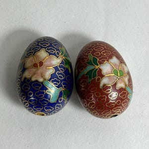 May include: Two decorative egg-shaped ornaments. One is dark blue with a floral design in pink, white, and gold. The other is red with a similar floral pattern. Both have gold outlines and are likely made of enamel.
