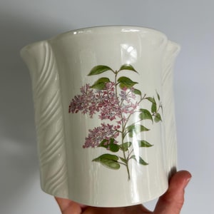 May include: Cream ceramic vase with a floral pattern. The vase features a lilac flower and green leaves. The sides have a textured, ridged design. The vase is held by a hand.