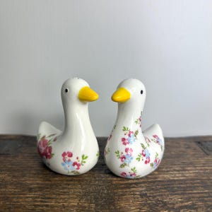 May include: Two white ceramic duck-shaped salt and pepper shakers. Each duck has a yellow beak, black eyes, and a floral pattern in pink and blue. The shakers are displayed on a wooden surface.
