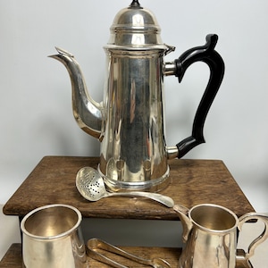 Puede incluir: Una cafetera plateada con asa negra y pomo en la tapa, acompañada de una cuchara plateada, dos jarras plateadas pequeñas y unas pinzas para azúcar. Los artículos se exhiben sobre una superficie de madera.