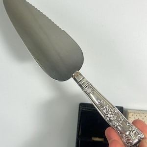 May include: Silver cake server with a serrated blade and an ornate handle. The handle features floral and scroll designs. The server is likely made of stainless steel or a similar metal. It is designed for cutting and serving cake.