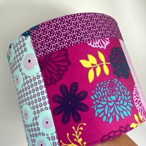 May include: A cylindrical lampshade with a vibrant floral pattern. The design features large magenta flowers, teal and yellow accents, and a geometric pattern at the top. The lampshade is held by a hand.