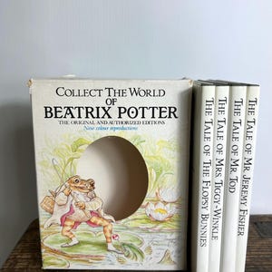 Peut inclure: Un coffret de livres de Beatrix Potter. La boîte présente une illustration de grenouille et le texte "Collect the World of Beatrix Potter". Quatre livres blancs avec des titres comme "The Tale of Mr. Jeremy Fisher" sont visibles.