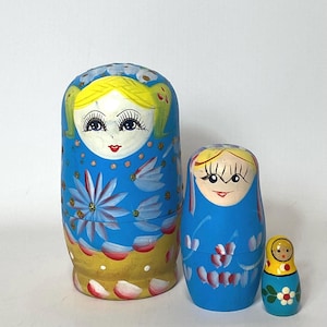 Peut inclure: Ensemble de trois poupées russes, également appelées Matriochkas, de tailles différentes. La plus grande poupée est bleue avec un motif floral et une base jaune. La poupée du milieu est également bleue avec un motif floral, et la plus petite est jaune et bleue.