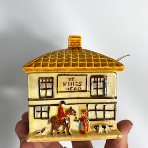 May include: A ceramic house-shaped container with a yellow tiled roof and the text "YE KINGS HEAD". The container features a scene with figures and dogs. A small spoon rests on the roof. The container is held by a hand.