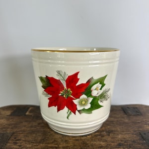 May include: A cream-coloured ceramic planter featuring a festive botanical illustration of a vibrant red poinsettia, white flowers, and green holly leaves. The cylindrical vessel has a subtle horizontal ribbed texture and a thin gold rim.