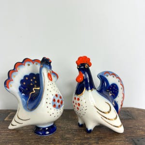 Puede incluir: Dos figuras de gallo de cerámica. Una es un jarrón con interior azul y cola en forma de abanico. La otra es un gallo con cuerpo blanco, detalles azules y rojos y ribete dorado. Ambas sobre una superficie de madera.