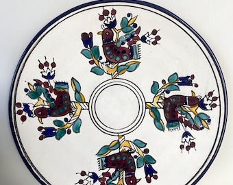May include: A white ceramic plate with a blue rim and a decorative design of four stylized birds in shades of red, brown, blue, and yellow.
