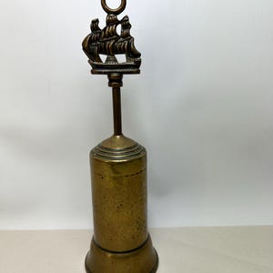 May include: A vintage brass bell featuring a ship design. The bell has a cylindrical body and a flared base. The ship is a detailed three-masted sailing vessel. The bell is likely made of brass and has a tarnished finish.