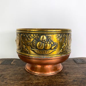 Peut inclure: Un bol décoratif en métal avec un bord doré et une base cuivrée. Le bol présente des motifs en relief de fruits et de feuillage. Le bol est posé sur une surface en bois.