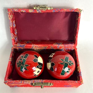 May include: A red, open box with two red, glossy balls inside. The balls have a bamboo design with black, white, and green accents. The box is lined with red fabric and has a gold-colored clasp.