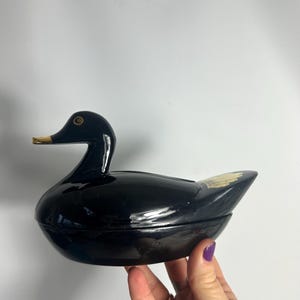 May include: A glossy black duck-shaped container with a golden beak and eye detail. The container has a lid and is held by a hand with purple nail polish. The duck has a decorative gold feather design on its back.