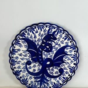 May include: A round ceramic plate with a scalloped edge, featuring a hand-painted dark blue dragon motif on a white background. The design includes intricate blue leaf-like patterns surrounding the central dragon figure and a dotted border.