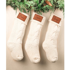 May include: Three white knitted Christmas stockings with brown leather name tags. The names on the tags are "Diana", "Sophia", and "George".