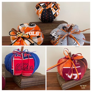 May include: Five handmade fabric pumpkins decorated with different college team logos. The pumpkins are orange, blue, red, and gray with black, white, and gold accents. The logos include Ole Miss, Virginia Tech, and the University of Tennessee. The pumpkins are sitting on a wooden surface.