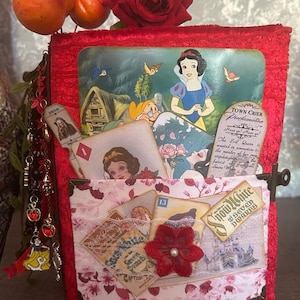 Peut inclure: Un journal rouge recouvert de tissu sur le thème de Blanche-Neige. Le journal présente une illustration de Blanche-Neige, des cartes à jouer et une poche remplie d'éphémères. Une rose rouge et des pommes factices ornent le dessus. Le journal a une fermeture à fermoir.