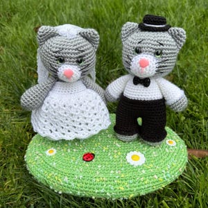 Könnte beinhalten: Ein gehäkelter Hochzeitstortenaufsatz mit einer grauen Katzenbraut in einem weißen Kleid und einem grauen Katzenbräutigam in einem schwarzen Smoking. Der Aufsatz steht auf einer grünen gehäkelten Basis mit weißen Blumen und einem roten Marienkäfer.