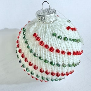 May include: White knitted Christmas ornament with red and green beads. The ornament is round and has a silver top.