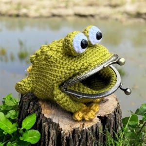 May include: A green crocheted frog coin purse with a wide-open mouth featuring a metallic, antique-style kiss-lock clasp. The frog has two large, white, protruding eyes with black pupils and small yellow feet at the base of its textured body.