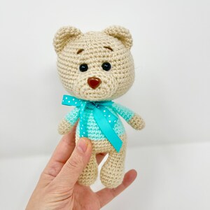 Könnte beinhalten: Ein gehäkelter Teddybär, der ein hellblaues Hemd mit weißen Punkten und eine türkisfarbene Schleife trägt. Der Bär hat braune Augen und eine braune Nase.