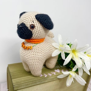 Puede incluir: Un juguete de perro pug de crochet con orejas y nariz negras, que lleva un collar naranja con una cuerda verde y cuentas de letras naranjas que deletrean "ABBEY". El juguete está sentado en una caja de madera con flores blancas.