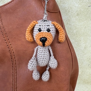 Puede incluir: Un llavero de perro de ganchillo gris con orejas naranjas, ojos negros y nariz negra. El llavero está unido a un bolso de cuero marrón.