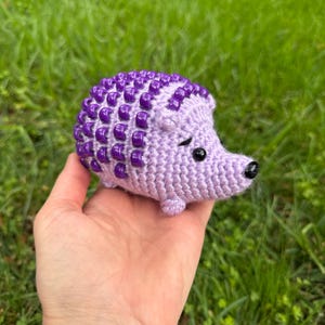 Könnte beinhalten: Ein handgehäkelter Igel in Lavendel- und Lilatönen. Der Körper ist aus weichem Lavendel, mit einem strukturierten Design aus lila Perlen auf dem Rücken. Der Igel hat kleine schwarze Augen und eine schwarze Nase. Das Spielzeug wird in einer Hand vor einem verschwommenen grünen Hintergrund gehalten.