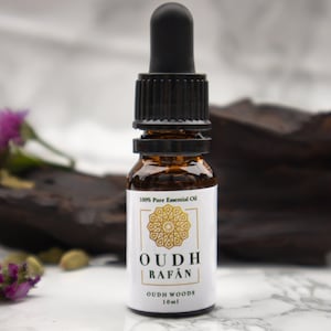 Puede incluir: Un pequeño frasco de vidrio marrón con un gotero negro. El frasco está etiquetado con "OUDH RAFAN" y "OUDH WOODS 10ml". El frasco está sobre una superficie blanca con un trozo de madera oscura y flores moradas.