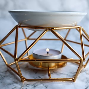 Può includere: Un supporto per diffusore geometrico in metallo dorato con una ciotola in ceramica bianca in cima e una piccola candela da tè bianca al centro.