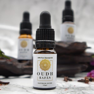 Puede incluir: Un pequeño frasco de vidrio marrón con un gotero negro. El frasco está etiquetado con "100% Pure Essential Oil" y "OUDH RAFAN SAFFRON OUDH 10ml". El frasco está sentado sobre un trozo de madera oscura con flores moradas y especias esparcidas a su alrededor.