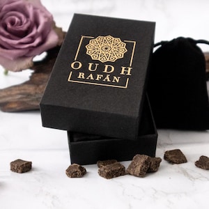May include: A black box with gold lettering that reads "OUDH RAFAN" is open, revealing a product. The box is next to a black velvet pouch, pieces of a product, and two purple roses. The background is white.