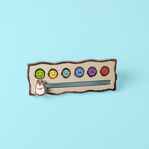 May include: A silver metal pin with a brown background and a row of seven colourful smiley faces. The faces range from happy to sad. A small brown cartoon character is on the left side of the pin.