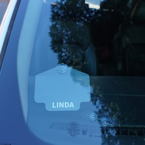 May include: A gray car windshield with a blue plastic tag that says "LINDA" on it.