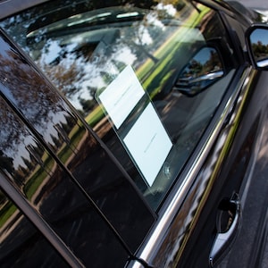 May include: A black car with a white paper notice on the back passenger window. The car is parked on a paved surface.