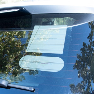 May include: A rear window of a car with a white paper note attached to the window with a suction cup. The note has lines and the text "Place your DMV sticker here".
