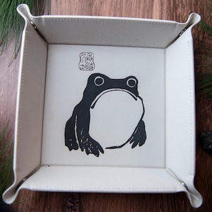 Pode incluir: Bandeja de couro sintético branco com uma ilustração de sapo em preto e branco. O sapo está centrado na base da bandeja, com um selo japonês acima. A bandeja tem cantos dobrados e está sobre uma superfície de madeira.