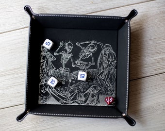 May include: A black leather dice tray with a white design of skeletons dancing. There are five dice inside the tray, four white dice with blue numbers and one red die with black numbers.