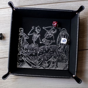 K&ouml;nnte beinhalten: Schwarzes Leder-W&uuml;rfeltablett mit einem wei&szlig;en gestickten Design von tanzenden Skeletten. Zwei W&uuml;rfel sind im Tablett sichtbar, einer rot und einer wei&szlig;.