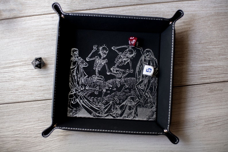 K&ouml;nnte beinhalten: Schwarzes Leder-W&uuml;rfeltablett mit einem wei&szlig;en gestickten Design von tanzenden Skeletten. Zwei W&uuml;rfel sind im Tablett sichtbar, einer rot und einer wei&szlig;.