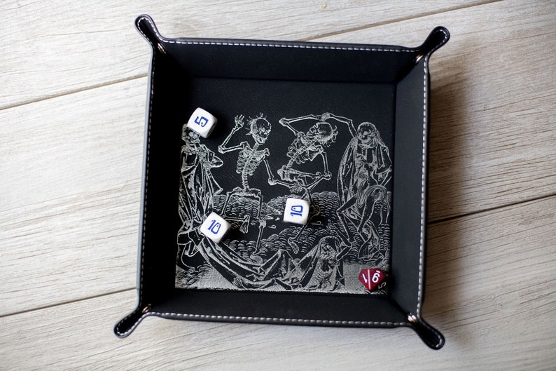K&ouml;nnte beinhalten: Ein schwarzes Leder-W&uuml;rfeltablett mit einem wei&szlig;en Design von tanzenden Skeletten. Es befinden sich f&uuml;nf W&uuml;rfel im Tablett, vier wei&szlig;e W&uuml;rfel mit blauen Zahlen und ein roter W&uuml;rfel mit schwarzen Zahlen.
