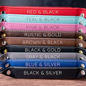 K&ouml;nnte beinhalten: Ein Stapel von neun Ledertabletts in verschiedenen Farben, darunter Rot, Teal, Pink, Braun, Schwarz, Grau und Blau. Jedes Tablett hat einen kontrastierenden Farbrahmen und einen Metallknopf an jeder Seite. Die Tabletts sind mit ihren Farbkombinationen beschriftet, wie z. B. "Rot & Schwarz", "Teal & Schwarz" und "Pink & Schwarz".