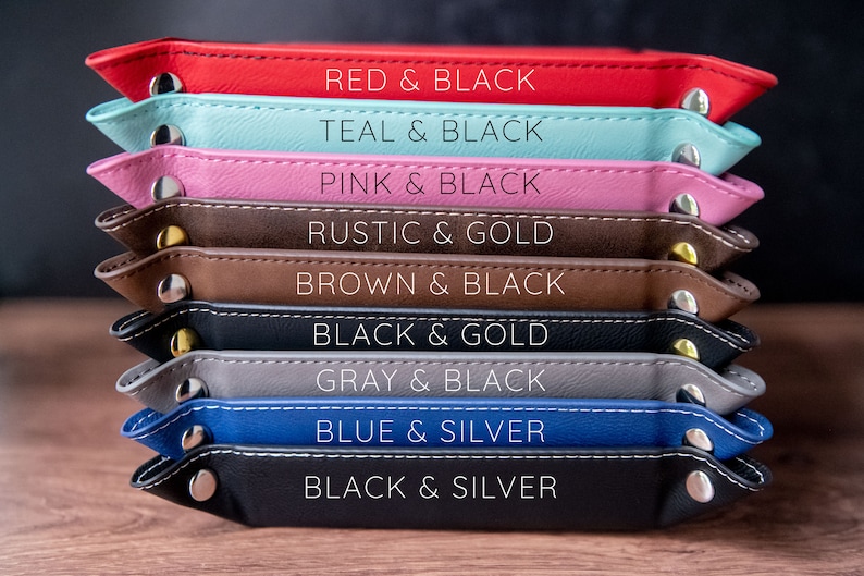 K&ouml;nnte beinhalten: Ein Stapel von neun Ledertabletts in verschiedenen Farben, darunter Rot, Teal, Pink, Braun, Schwarz, Grau und Blau. Jedes Tablett hat einen kontrastierenden Farbrahmen und einen Metallknopf an jeder Seite. Die Tabletts sind mit ihren Farbkombinationen beschriftet, wie z. B. "Rot & Schwarz", "Teal & Schwarz" und "Pink & Schwarz".